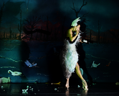 A ballerina dressed in a swan costume lights a cigarette on a dark stage.