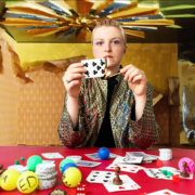 HouseNeverWins1 :: A female with blonde hair pinned back sits centre, leaning on a red poker table covered in cards, poker chips and ping pong balls. She is burning the 9 of clubs with a match. In the background is a colourful casino with the wallpaper peeling and falling apart.